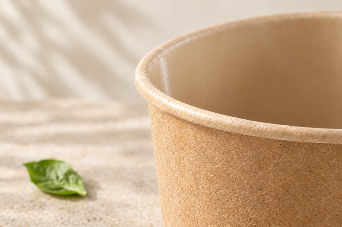 Close-up of the rim and inner wall of a kraft paper cup, showing the paper fibres and the smooth inner lining, with a green leaf beside it.